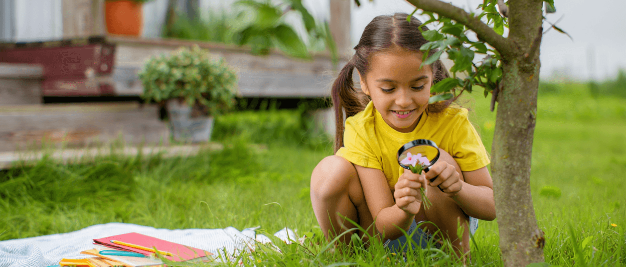 Child with pigtails examining pink flowers with a magnifying glass.